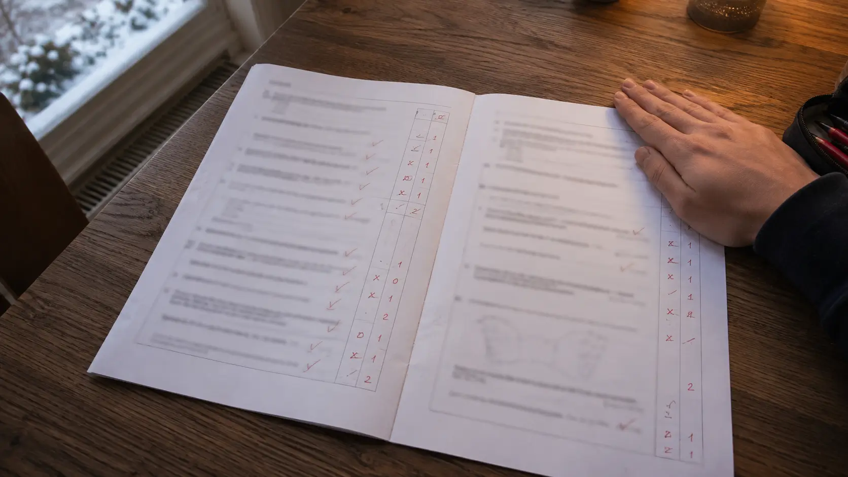 A marked mock exam paper open on a table in winter, red ticks and marks visible question by question, a hand resting where the grade would be