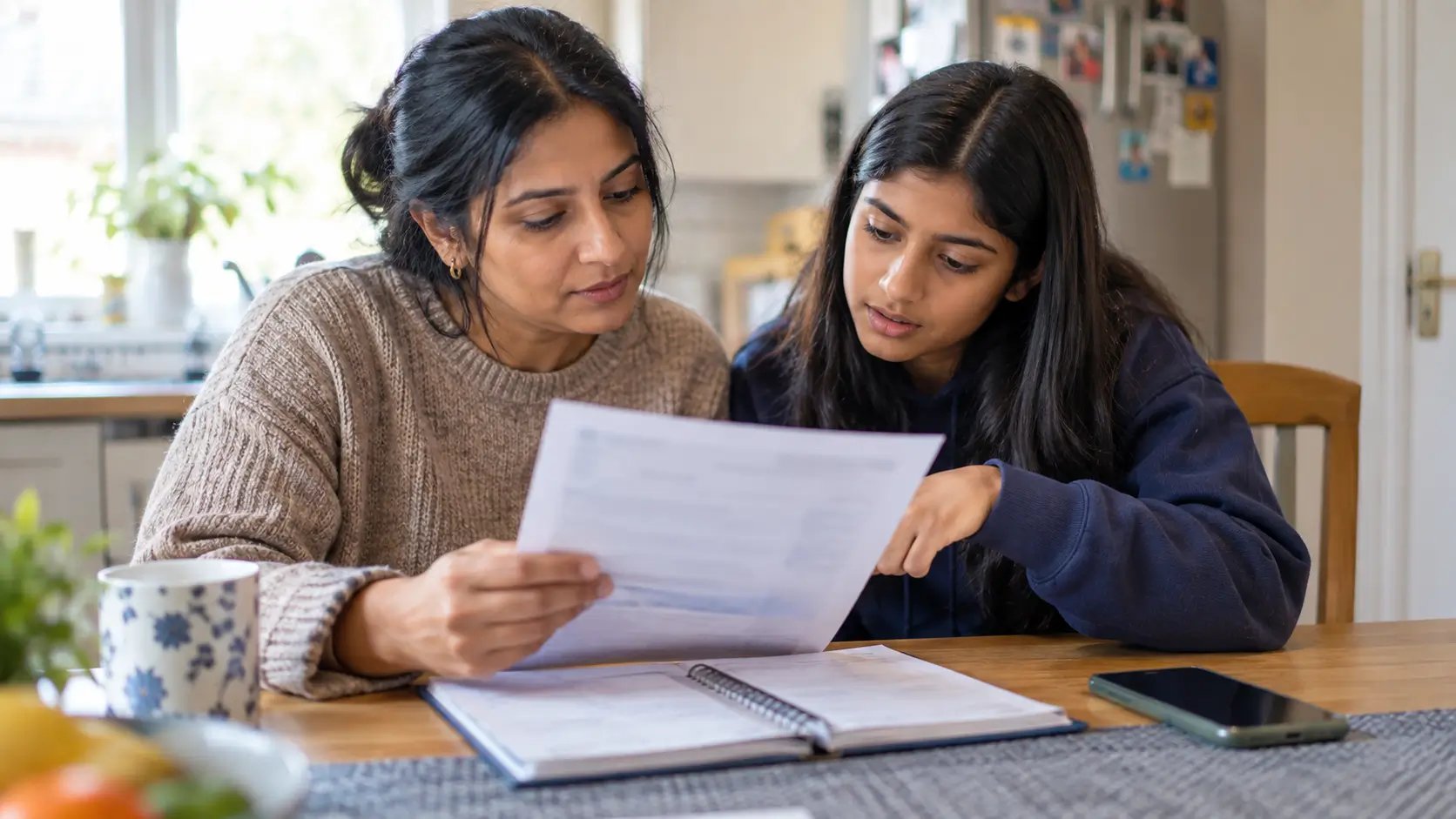 A mother and her teenage daughter reading a school letter together at their kitchen table, the daughter pointing at a line on the page